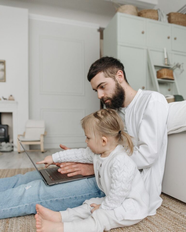 Father working on a laptop at home with his young daughter beside him